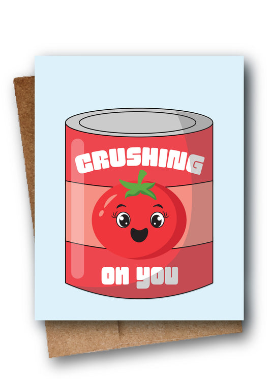 A square greeting card with a light blue background featuring a cartoon red tomato can labeled 'CRUSHING on you,' shown leaning against a brown kraft envelope.