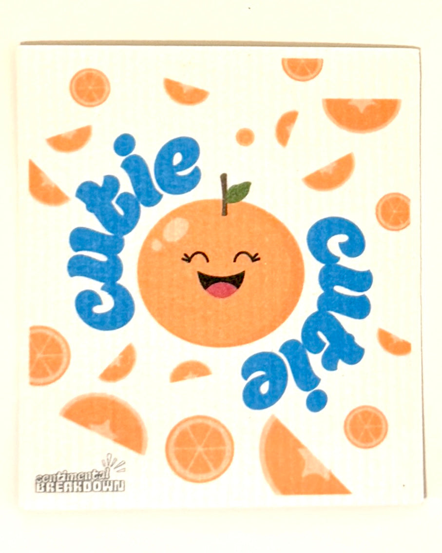 A square white paper napkin with a waffle texture, printed with a cute orange face, orange slices, and the word 'cutie' in blue script, displayed on a plain light background.