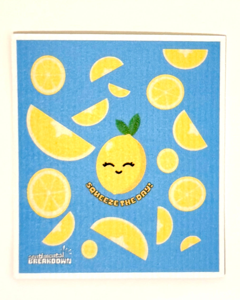 A square kitchen towel with a blue background and a cute yellow lemon illustration, displayed flat against a neutral light beige background.