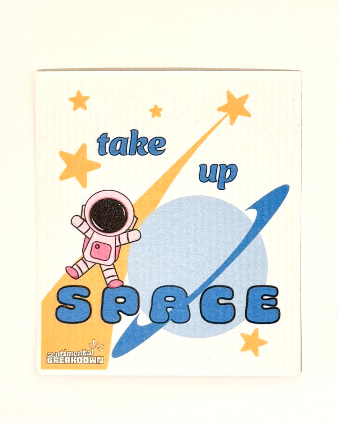 A square white paper napkin with a colorful space-themed graphic that reads 'take up SPACE' in blue and yellow, featuring an astronaut, stars, and a planet, photographed on a plain off-white surface.