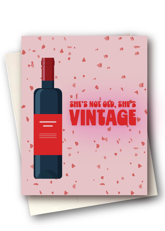 A pink greeting card with a red wine bottle illustration and the text 'SHE'S NOT OLD, SHE'S VINTAGE' in red, shown with cream envelopes behind it on a white background.