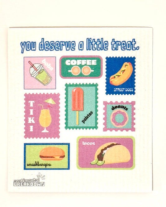 A white textured paper napkin printed with colorful food illustrations and the phrase 'you deserve a little treat.' in blue text, photographed against a plain light background.