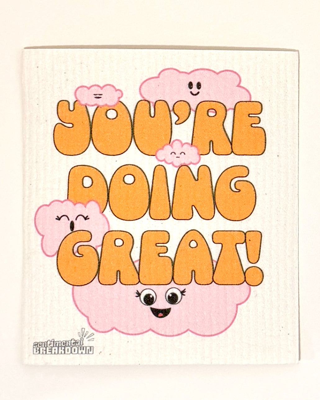 A square decorative dish towel or napkin with a white ribbed background, featuring the phrase 'YOU'RE DOING GREAT!' in bold orange letters, surrounded by cute pink smiling cartoon clouds.